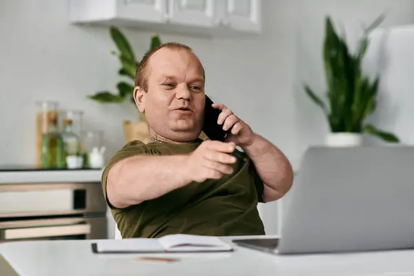 Un homme avec inclusivité sur un appel téléphonique dans son bureau à la maison, assis à un bureau avec un ordinateur portable et un bloc-notes. — Photo de stock