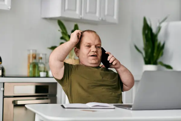 A man with inclusivity sits at his home office desk, talking on the phone, with a laptop and a notebook in front of him. — Stock Photo