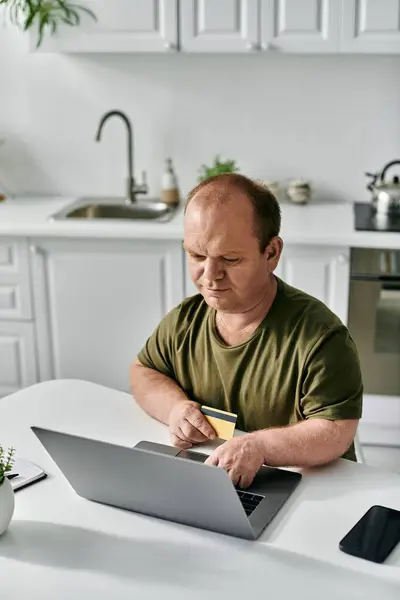 A man with inclusivity sits at a kitchen table, using his laptop and a credit card for online shopping. — Stock Photo