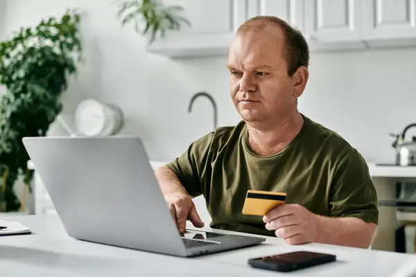 Um homem com inclusão senta-se em uma mesa em sua casa, usando um laptop e um cartão de crédito para fazer uma compra. — Fotografia de Stock