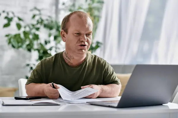 Un hombre con inclusividad se sienta en un escritorio en una oficina en casa, revisando documentos mientras mira una computadora portátil. - foto de stock