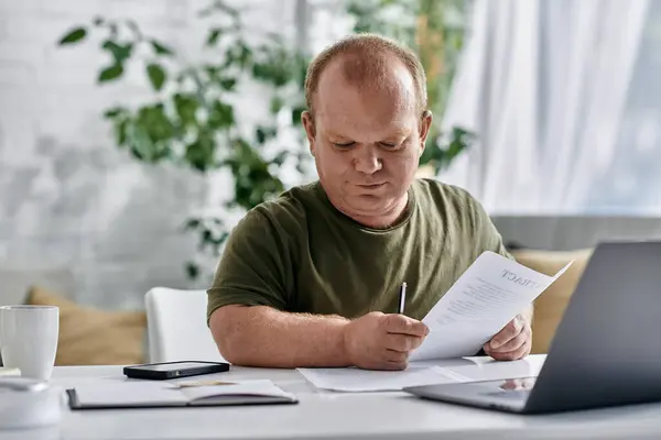 A man with inclusivity sits at a table, reviewing paperwork while working from home. — Stock Photo
