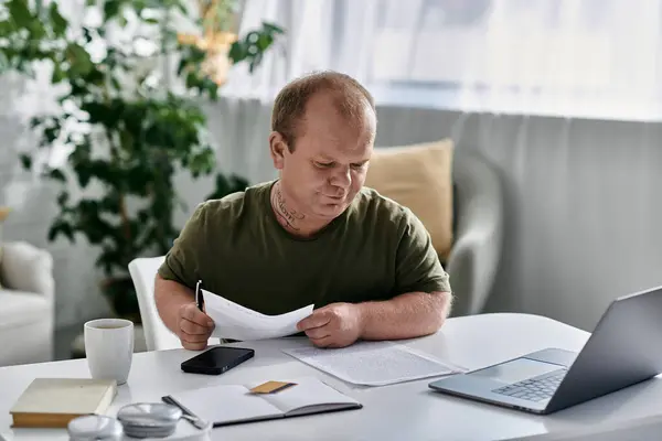 A man with inclusivity sits at a table in his home office, reviewing paperwork while a laptop sits on the table. - foto de stock