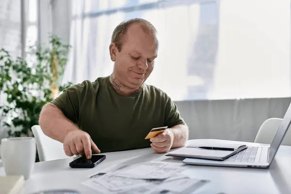 Un uomo con inclusività in abbigliamento casual usa il telefono e la carta di credito mentre è seduto presso una scrivania di un home office. — Foto stock