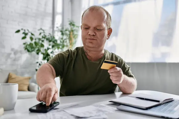 Un homme avec inclusivité est assis à une table dans sa maison, effectuant un paiement avec une carte de crédit. — Photo de stock