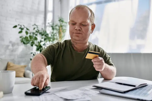 Un homme avec inclusivité est assis à une table dans sa maison, effectuant un paiement avec une carte de crédit. — Photo de stock