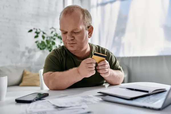 Человек с инклюзивностью сидит за столом в своем доме, держит кредитную карту и внимательно на нее смотрит.. — стоковое фото
