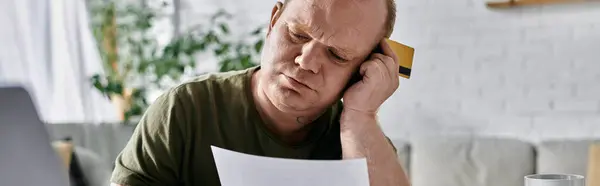 Un uomo con inclusività si siede a casa, apparentemente gravato da preoccupazioni finanziarie, mentre esamina un documento e detiene una carta di credito.. — Foto stock