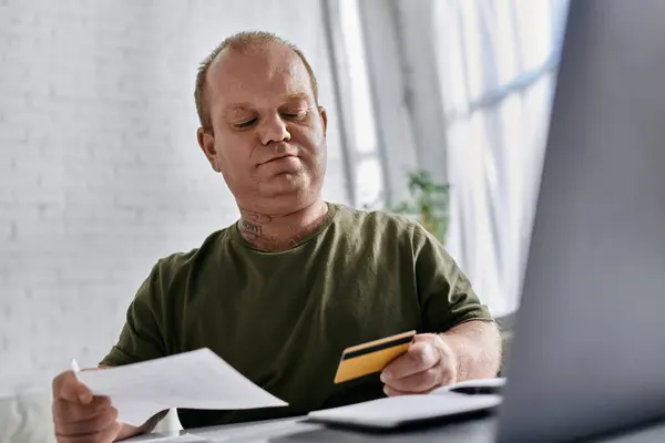 Un uomo con inclusività in abbigliamento casual si siede a una scrivania, rivedere le bollette e una carta di credito. — Foto stock