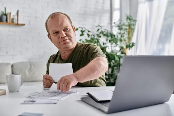 Человек с инклюзивностью сидит за своим домашним столом, просматривает документы и работает над своим ноутбуком. — стоковое фото