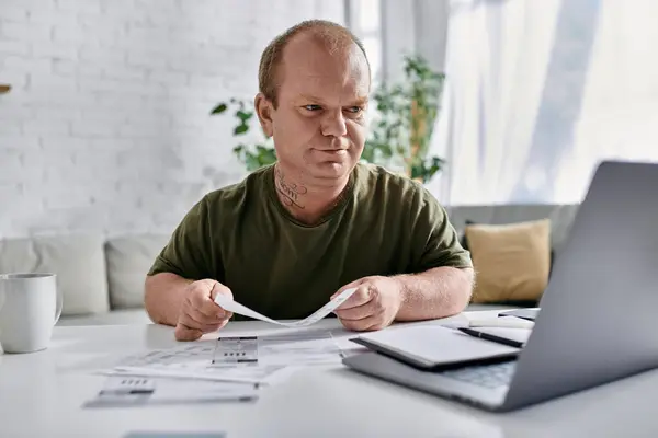 Un hombre con inclusividad revisa los papeles en su escritorio de casa con un portátil abierto delante de él. - foto de stock