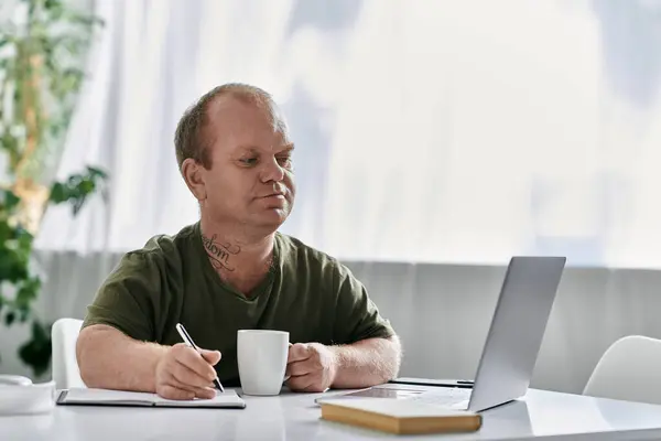Un uomo con inclusività siede a un tavolo nella sua casa, vestito con disinvoltura, scrivendo in un quaderno mentre usa un computer portatile. — Foto stock