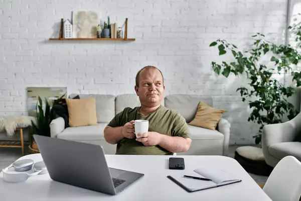 A man with inclusivity sits at a desk in a casual home office setting, holding a mug and gazing thoughtfully into the distance. — Stock Photo