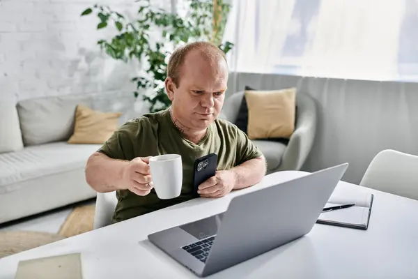 Un homme avec inclusivité en tenue décontractée s'assoit à une table dans sa maison, dégustant une tasse de café tout en utilisant un ordinateur portable et un smartphone. — Photo de stock