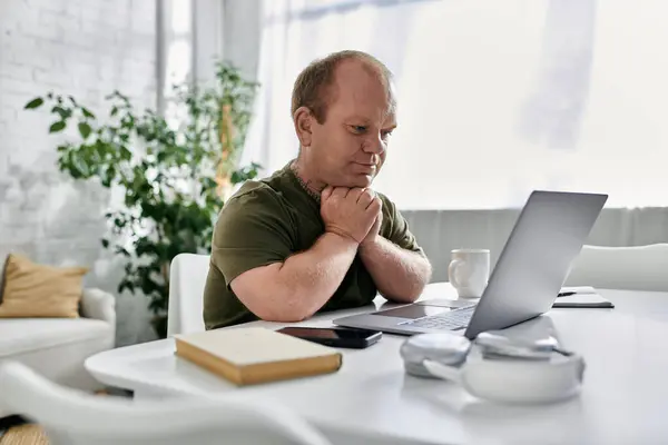 A man with inclusivity in casual clothing is sitting at a table in his home, working on his laptop. — Stock Photo