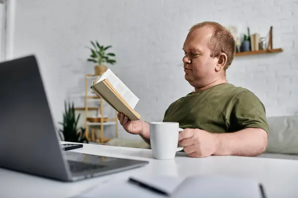 Человек с инклюзивностью сидит за столом в своем доме, читает книгу и держит чашку кофе. — стоковое фото