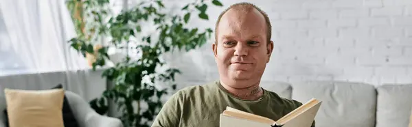 A man with inclusivity relaxes on a couch, casually dressed, reading a book. - foto de stock