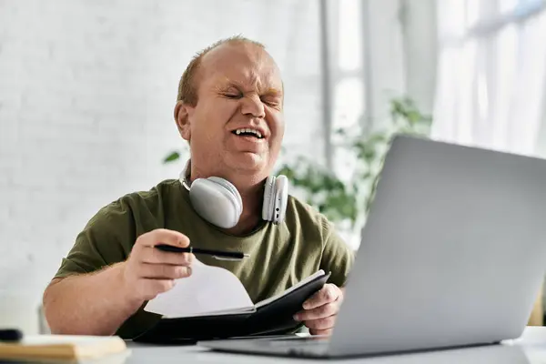 A man with inclusivity with headphones sits at his desk working on a laptop and taking notes. — Stockfoto