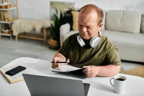 Ein Mann mit Inklusion sitzt zu Hause an einem Tisch, trägt Kopfhörer und schreibt in ein Notizbuch. — Stockfoto