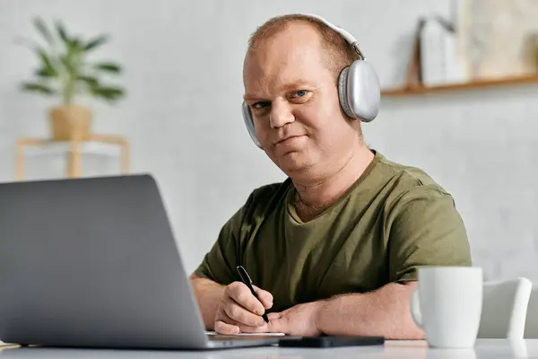 A man with inclusivity wearing headphones sits at a table, using a laptop and taking notes. — Stock Photo