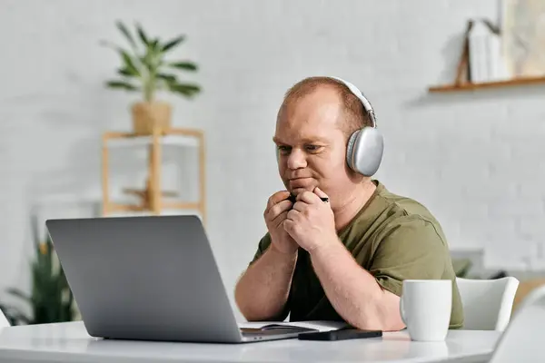 Un homme avec inclusivité est assis à un bureau dans son bureau à la maison, travaillant sur son ordinateur portable tout en portant des écouteurs. — Photo de stock