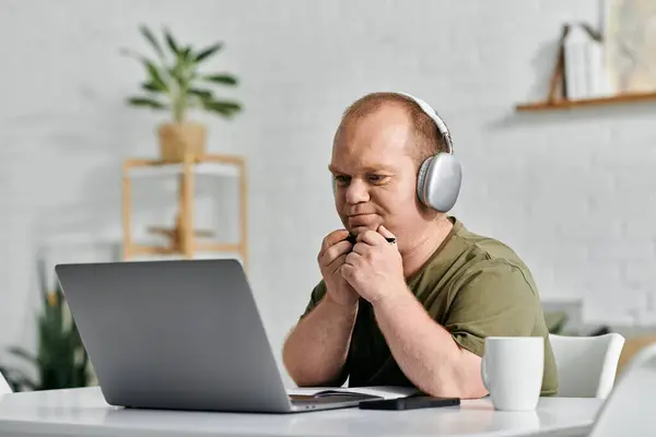 A man with inclusivity sits at a desk in his home office, working on his laptop while wearing headphones. — Stock Photo