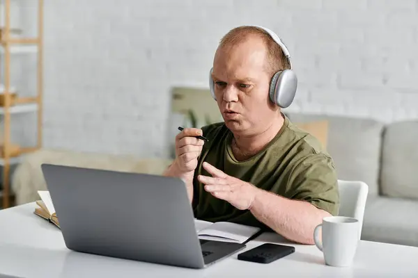 A man with inclusivity in casual clothing sits at a desk, wearing headphones and using a laptop. — Stock Photo