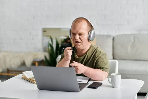 A man with inclusivity in casual attire sits at a desk in his home, using a laptop and headphones to engage in a passionate online conversation. — Stock Photo