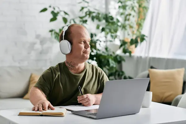 A man with inclusivity wearing headphones sits at a desk with a laptop, a book, and a pen, working from home. — Stock Photo