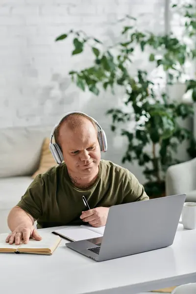 Ein Mann in Freizeitkleidung und mit Kopfhörern sitzt an einem Schreibtisch mit Laptop und aufgeschlagenem Buch und konzentriert sich auf seine Arbeit.. — Stockfoto