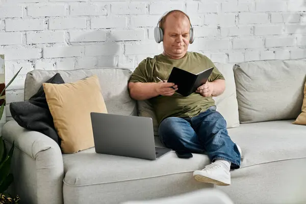 A man with inclusivity sits on a sofa at home, wearing headphones and writing in a notebook. - foto de stock