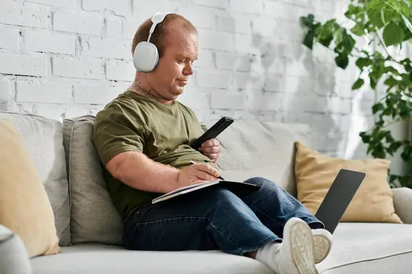 A man with inclusivity wearing headphones sits on a couch at home, comfortably relaxed while using a laptop and taking notes. — Stock Photo