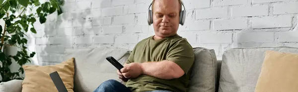 A man with inclusivity sits comfortably on a couch, wearing headphones and holding a smartphone. — Stockfoto