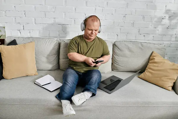 A man with inclusivity relaxes on a couch at home, wearing headphones and checking his phone. — Stock Photo