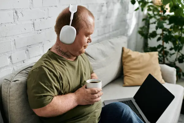 Ein Mann mit Inklusivität sitzt zu Hause auf einer Couch, trägt Kopfhörer, genießt eine Tasse Kaffee und benutzt einen Laptop. — Stockfoto