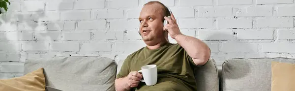 A man with inclusivity wearing headphones and casual attire relaxes on a couch at home, holding a cup of coffee. — Stock Photo