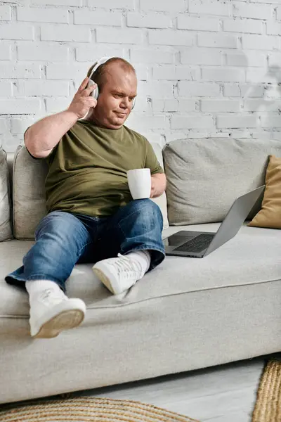 A man with inclusivity wearing headphones and casual clothing sits on a couch at home, enjoying a relaxing moment with a laptop and a cup. — Stock Photo