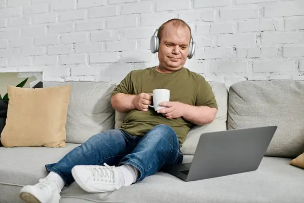 Un homme avec inclusivité se détend sur un canapé à la maison, portant un casque et tenant une tasse de café. — Photo de stock