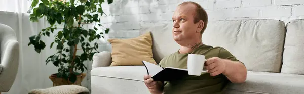 A man with inclusivity is sitting on a couch in his living room, reading a book and enjoying a cup of coffee. — Stock Photo