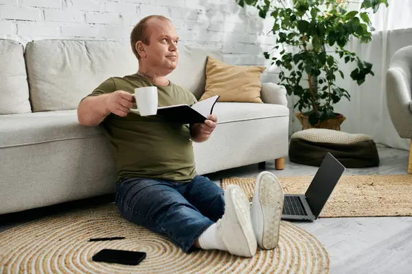 A man with inclusivity sits on the floor in a living room, enjoying a cup of coffee and a book. - foto de stock