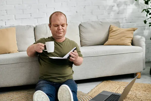 A man with inclusivity relaxes on the floor at home, enjoying a cup of coffee and a book. - foto de stock