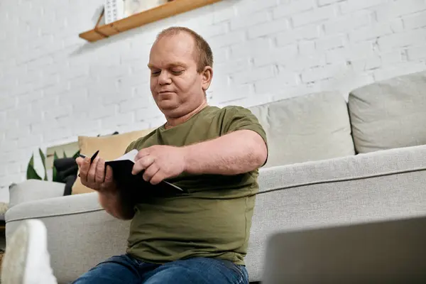A man with inclusivity wearing a green t-shirt and jeans sits on a couch reading a book. - foto de stock