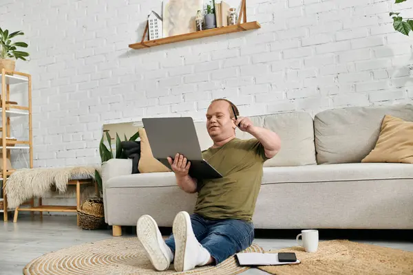 A man with inclusivity sits on the floor in front of a couch, working on a laptop. — Stock Photo