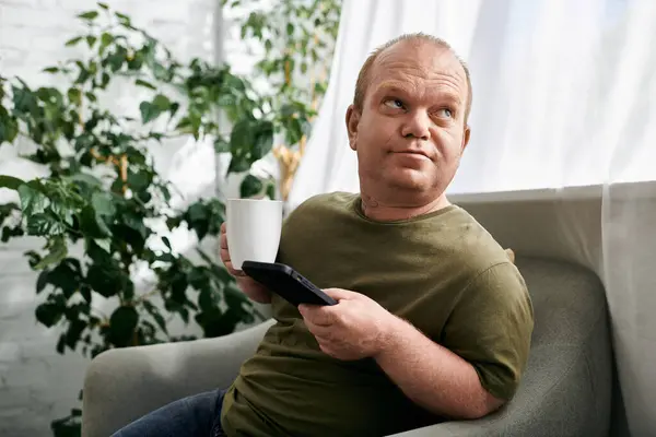 Un homme avec inclusivité est assis sur une chaise, tenant une tasse de café et levant les yeux pensivement. — Photo de stock