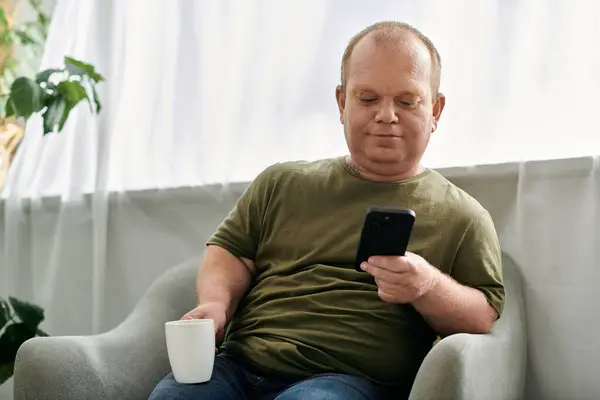 Un homme avec inclusivité se détend dans une chaise à la maison, dégustant une tasse de café et parcourant son téléphone. — Photo de stock