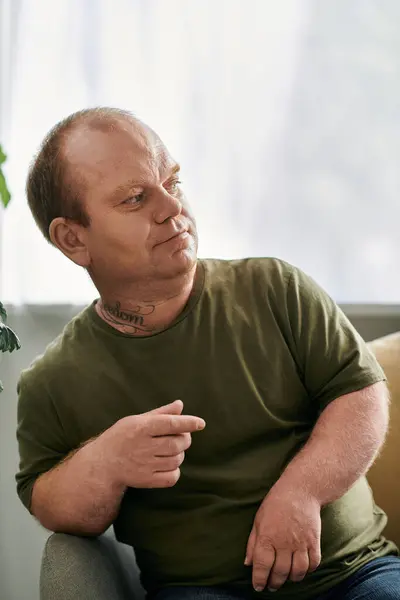 Un uomo con inclusività si siede comodamente nella sua casa, vestito con disinvoltura, guardando pensieroso di lato. — Foto stock