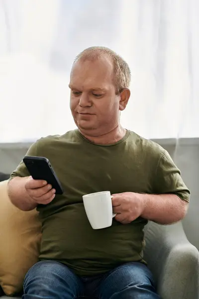 Un homme avec inclusivité en tenue décontractée s'assoit sur un canapé, tenant une tasse de café dans une main et un smartphone dans l'autre. — Photo de stock