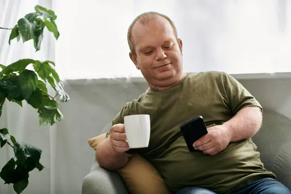 Un homme avec inclusivité est assis dans une chaise confortable à la maison, profitant d'une tasse de café tout en regardant son téléphone. — Photo de stock