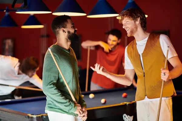 Gli amici stanno accanto a un tavolo da biliardo, parlando e ridendo mentre il loro amico gioca un colpo sullo sfondo. — Foto stock