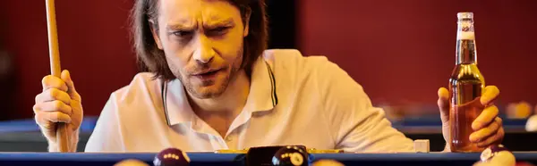 Un uomo tiene un bastone mentre gioca a biliardo con gli amici, con una bottiglia di birra in primo piano. — Foto stock